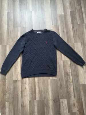 Mörkblå tröja från Polo Ralph Lauren - Mörkblå tröja från Polo Ralph Lauren med rutmönstrad framsida och röd broderad logga på bröstet. Tröjan har rund halsringning, långa ärmar och ribbade muddar. Perfekt för dig som gillar klassisk och stilren design. Storlek M