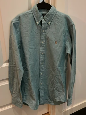 Ljusblå skjorta från Ralph Lauren - Klassisk blå-grön skjorta från Ralph Lauren med button-down krage och broderad logga på bröstet. Skjortan har långa ärmar och knäppning framtill. Perfekt till jeans eller chinos för en clean och stilren look.