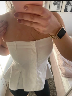 Peplumtopp/Festopp Vit axelbandslös korsettopp - Snygg vit axelbandslös korsettopp med peplumdetalj och markerad midja. Toppen har en strukturerad form och stängs framtill. Perfekt att styla med jeans eller kjol för en trendig look. Har en osynlig dragkedja på sidan och knappar framtill, så enkel att ta på  passar S/M