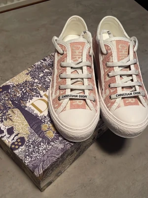 Christian Dior rosa låga sneakers - Säljer ett par snygga låga sneakers från Christian Dior i rosa och vitt med tydligt Dior-mönster. Skorna har vita snören, gummisula och detaljer med logotypen på tungan och snörningen. Perfekta för dig som vill ha något exklusivt och trendigt.