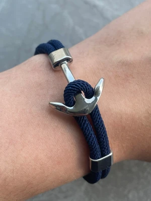 Marint armband med ankardetalj - Snyggt armband med marinblått rep och en silverfärgad ankare i metall som lås. Armbandet har en modern och stilren design med metalliska detaljer som ger en cool nautisk känsla. Perfekt accessoar för dig som gillar havsinspirerad stil.