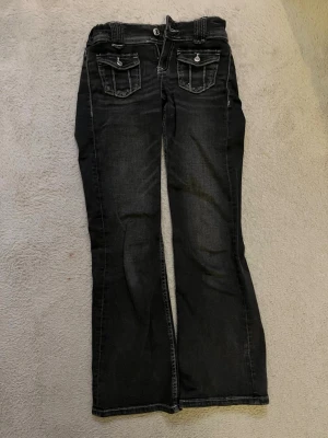 Svarta bootcut jeans med fickdetaljer - Använd gärna köp nu!!🩷 Säljer ett par svarta bootcut jeans med snygga fickdetaljer och kontrastsömmar. Jeansen har två fickor fram med lock och knapp, samt bälteshällor. Perfekta för dig som gillar en avslappnad men trendig look.