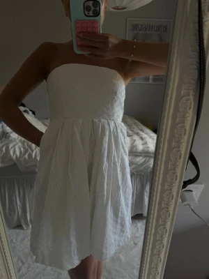 Vit axelbandslös kort klänning - Säljer en söt vit axelbandslös klänning med broderade detaljer och utställd kjol. Klänningen har en kort längd och är perfekt för varma dagar. Materialet känns lätt och luftigt, och modellen ger en fräsch och somrig look.