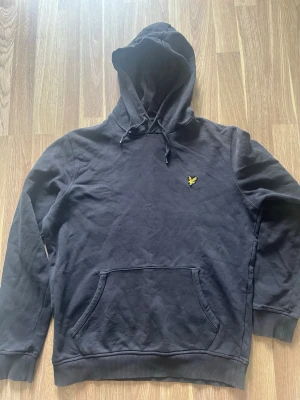 Svart hoodie från Lyle & Scott - Snygg svart hoodie från Lyle & Scott med klassisk gul logga på bröstet. Hoodien har dragsko i huvan och en stor magficka framtill. Tillverkad i mjukt bomullsmaterial och har ribbade muddar vid ärmslut och nederkant. Perfekt för en avslappnad stil.