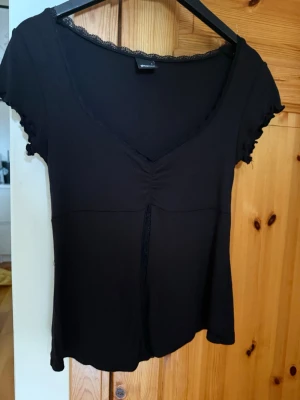 Svart topp med spets från Gina Tricot - Snygg svart topp från Gina Tricot med v-ringning, spetsdetaljer vid halsen och korta ärmar. Toppen har en lätt rynkning framtill och är tillverkad i mjukt material som sitter bekvämt. Perfekt att styla med jeans eller kjol.