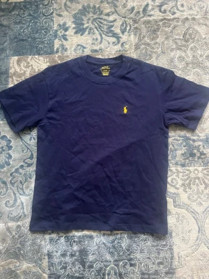 Marinblå t-shirt från Polo Ralph Lauren storlek M - Säljer en marinblå t-shirt från Polo Ralph Lauren med klassisk gul logga broderad på bröstet. T-shirten har rund hals och korta ärmar, perfekt för en avslappnad och stilren look. Materialet är mjuk bomull som känns skönt mot huden. T-shirten är helt ny och aldrig buren. Storlek M