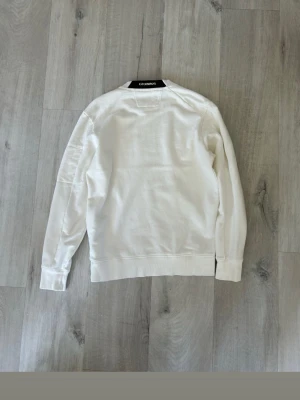 Vit sweatshirt från C.P. Company - Säljer en stilren vit sweatshirt från C.P. Company med rund halsringning och ribbade muddar. Tröjan har en ikonisk svart patch och ficka med dragkedja på ena ärmen. Perfekt för dig som gillar streetwear och enkel design.