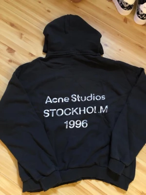 Svart hoodie Acne Studios Stockholm - Säljer en svart hoodie från Acne Studios med stor vit text på ryggen: 'Acne Studios STOCKHOLM 1996'. Klassisk passform med huva och ribbade muddar. Perfekt för dig som gillar streetwear och vill ha något från ett ikoniskt svenskt märke.