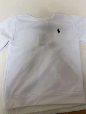 Vit sweatshirt från Ralph Lauren - Klassisk vit sweatshirt från Ralph Lauren med rund halsringning och den ikoniska svarta logotypen broderad på bröstet. Tröjan har långa ärmar och ribbade muddar vid ärmslut och nederkant. Perfekt för en clean och stilren look.