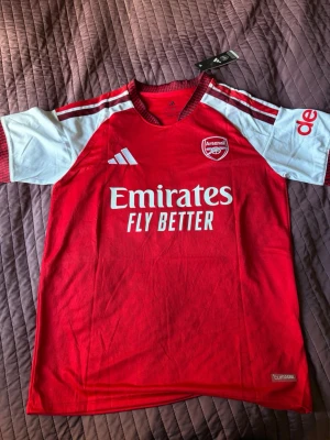 Arsenal matchtröja röd/vit Adidas - Säljer en officiell Arsenal matchtröja från Adidas i rött och vitt med tryck 'Emirates Fly Better' på bröstet och 'GYÖKERES 14' på ryggen. Tröjan har korta ärmar, klubbmärke på bröstet och är tillverkad i lätt funktionsmaterial.