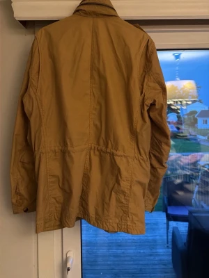 Beige utilityjacka med fickor från peak performance  - Snygg beige utilityjacka med fyra stora fickor framtill och axelklaffar. Jackan har hög krage och dragsko i midjan för en schysst passform. Perfekt för lager på lager och streetwear-vibe. Tillverkad i ett lätt bomullsliknande material.