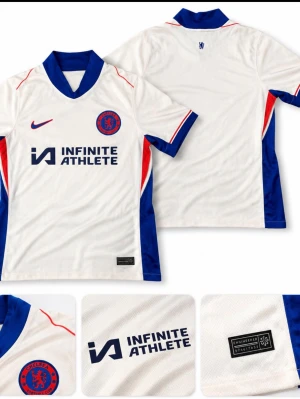Chelsea FC vit matchtröja Nike - Chelsea FC:s officiella matchtröja i vitt med blå krage och blå/röda detaljer längs sidorna. Tröjan har klubbens logga på bröstet, Nike-logga och sponsortryck 'Infinite Athlete'. Materialet är lätt och andas, perfekt för sport och träning.