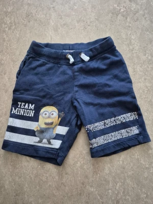 Shorts - Mörkblå shorts med tryck av en Minion och texten 'TEAM MINION' på vänster ben. Shortsen har vita och blå ränder, resårmidja med snörning och är gjorda i mjuk bomull. Perfekta för sommardagar och chill. Färgen har gått av lite.