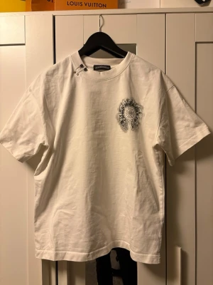 Vit t-shirt från Chrome Hearts - Vit t-shirt från Chrome Hearts med svart tryck på bröstet och två säkerhetsnålar vid halsen som cool detalj. Klassisk passform och rund halsringning. Perfekt för dig som gillar streetwear och unika detaljer. Fråga om du har någon fundering