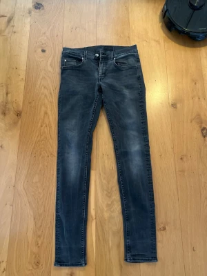 Slim fit svarta jeans Tiger of Sweden 32/34 - Slim fit svarta jeans i modellen Raleigh, tillverkade i Italien. Jeansen har klassisk femficksdesign, knappgylf och diskreta slitningar för en snygg tvättad look. Perfekta för dig som gillar en smal passform och stilren design.