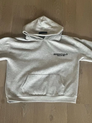 Grå Essentials Fear of God hoodie - Snygg ljusgrå hoodie från Essentials Fear of God med svart logga tryckt på bröstet och ryggen. Tillverkad i mjukt bomullsmaterial som känns skönt mot huden. Perfekt för en avslappnad streetwear-look.