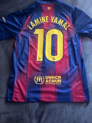  Lamine Yamal #10 Barcelona matchtröja - Säljer en FC Barcelona matchtröja med Lamine Yamal och nummer 10 på ryggen. Tröjan är blå och röd med gula detaljer, korta ärmar och klubbmärke på bröstet. Materialet är lätt och andas, perfekt för fotboll eller träning.