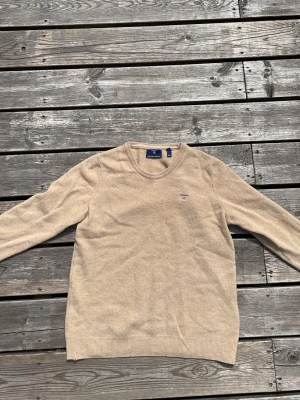 Beige stickad tröja från Gant - Snygg beige stickad tröja från Gant med rund halsringning och diskret logga broderad på bröstet. Tröjan har långa ärmar och ribbade muddar vid ärmslut och nederkant. Perfekt för lager på lager och enkel att matcha.