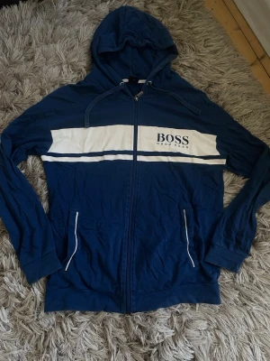 Blå hoodie från Hugo Boss - Snygg blå hoodie från Hugo Boss med vit panel och logga på bröstet. Dragkedja framtill, huva med snören och två fickor med vita detaljer. Materialet är mjukt och bekvämt, perfekt för en avslappnad look.