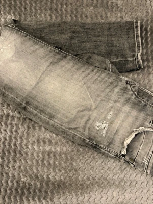 Grå slitna jeans med hål - Säljer ett par grå jeans med slitningar och hål vid knät. Jeansen har en tvättad look och detaljerade bakfickor. Perfekta för en avslappnad och trendig stil. Slim/glenn, 28/32
