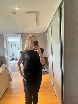 Svart volangblus med axelvolanger - Snygg svart blus med volangdetaljer vid axlarna och lös passform. Perfekt till jeans eller kjol för en trendig look. Blusen är ärmlös och har en enkel, stilren design utan synliga knappar eller dragkedja.                                                                           