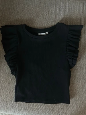 Svart volangtopp från Zara - Snygg svart topp från Zara med volangdetaljer längs axlarna. Toppen är ribbad och har en croppad passform som ger en trendig look. 