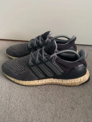Adidas Ultra Boost - Grå Adidas Ultra Boost sneakers med stickad ovandel i mesh för skön andningsförmåga. Skon har de klassiska tre ränderna på sidan och en vit/gulaktig Boost-sula som ger grym komfort. Perfekt för dig som gillar stilrena och sportiga sneakers.