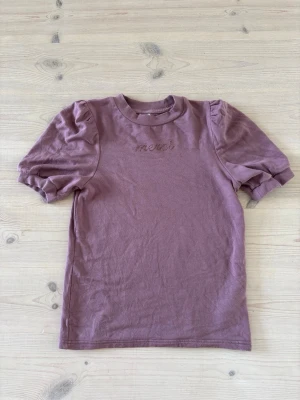 Ljuslila t-shirt med puffärm - Trendig ljuslila t-shirt med puffärmar och broderad text 'merci' framtill. Rund halsringning och mjukt bomullsmaterial gör den både bekväm och snygg. Perfekt för dig som vill ha en enkel men ändå unik t-shirt.