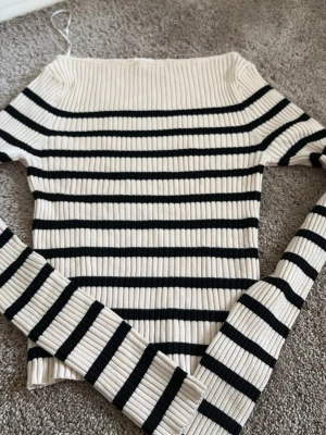 Randig ribbad tröja  - Trendig ribbad tröja med svarta och vita horisontella ränder och off shoulder-skärning. Långärmad modell i stretchigt material som sitter snyggt på kroppen. Perfekt för dig som gillar en stilren men ändå cool look.