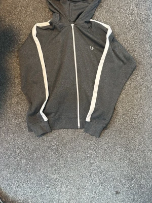 Mörkgrå hoodie från Fred Perry - Snygg mörkgrå hoodie från Fred Perry med vit dragkedja och vita ränder längs ärmarna. Klassisk huva och ribbade muddar. Perfekt för en avslappnad och sportig stil. Liten Fred Perry-logga på bröstet.xs- S