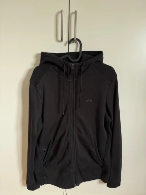 Svart hoodie med dragkedja från BOSS - Svart hoodie från BOSS med dragkedja framtill, huva med dragsko och två fickor. Diskret BOSS-logga på bröstet. Tillverkad i mjukt material som passar perfekt för en avslappnad stil.