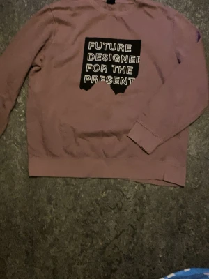 Lila sweatshirt från H&M - Säljer en lila sweatshirt med ett stort svart tryck på bröstet där det står 'FUTURE DESIGNED FOR THE PRESENT' i vita bokstäver. Tröjan har rund hals, långa ärmar och ribbade muddar. Perfekt för dig som gillar streetwear och statement-plagg.