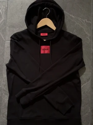 Svart hoodie från Hugo Boss - Säljer en svart hoodie från Hugo Boss med en röd logotyp på bröstet. Hoodien har huva med dragsnören och långa ärmar. Materialet är mjukt och bekvämt, perfekt för en avslappnad stil. Passar dig som vill ha en enkel men snygg look. Tröjan är väldigt bra skick, inga defekter. 