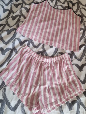 Lounge set - Säljer ett sött lounge-set med linne och shorts i rosa och vita ränder. Linnet har tunna svarta axelband och raka linjer, medan shortsen har resår i midjan för skön passform. Perfekt för mysiga kvällar hemma. Enbart testad