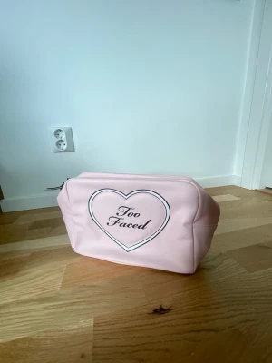 Rosa necessär från Too Faced - Söt rosa necessär från Too Faced i konstläder med hjärtdetalj och logga på framsidan. Väskan har dragkedja och ett randigt plast innerfoder i svartvitt, så den är lätt att torka av. Perfekt för smink eller småprylar när du är på språng. Oanvänd 