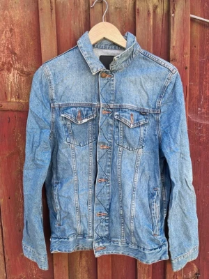 Klassisk blå jeansjacka - Flux Melbourne jeansjacka i klassisk modell och snygg blå tvätt. Jackan har två bröstfickor med lock och knappar, praktiska sidofickor, kontrastsömmar och metallknappar framtill.  Perfekt att bära över en hoodie eller t-shirt för en avslappnad och tidlös look.  Bra skick.