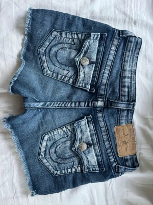 Blå jeansshorts från True Religion - Säljer ett par blå jeansshorts från True Religion med råa, fransiga benslut och klassiska kontrastsömmar. Shortsen har stora bakfickor med lock och silverfärgade knappar samt en läderpatch bak i midjan. Perfekta för sommaren!