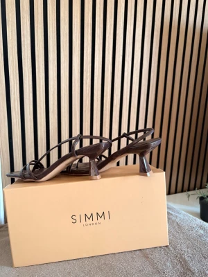 Bruna strappy pumps med krokopräglat mönster - Säljer ett par bruna pumps med smala remmar och fyrkantig tå. Skorna har ett krokopräglat mönster och en låg, vinklad klack. Materialet ser ut som konstläder och designen är minimalistisk men trendig. Perfekta för dig som gillar stilrena detaljer.