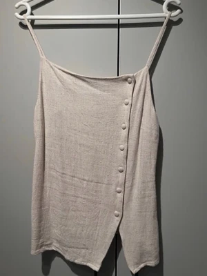 Beige linnetopp med knappar - Säljer en stilren beige linnetopp med smala axelband och dekorativa knappar framtill. Toppen har en lätt asymmetrisk skärning och är tillverkad i ett luftigt material som passar perfekt till varma dagar.
