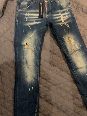 Blå slitna jeans med dragkedjedetalj - Säljer ett par blå jeans med slitningar, hål och färgstänk för en riktigt trendig look. Jeansen har skinny fit och en cool dragkedjedetalj vid gylfen. Perfekta för dig som gillar streetstyle och vill sticka ut.