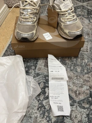 Beige och silver Asics sneakers - Snygga sneakers från Asics i beige och silver med mesh- och syntetmaterial. Skorna har klassisk låg profil, vita snören och tydliga Asics-loggor på sidan. Perfekta för dig som gillar sportig och trendig stil. Ny pris 1790kr