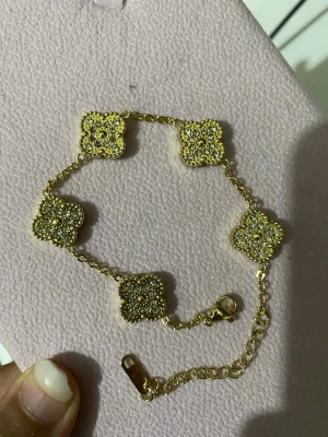Guldpläterad armband med pärldetaljer 18+5 - Snyggt armband i guldfärg med fem fyrklöverformade länkar, dekorerade med små glittrande pärlor. Kedjan har justerbar längd och ett smidigt lås. Perfekt accessoar för att lyfta din outfit med lite extra bling.
