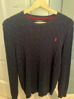 Mörkblå kabelstickad tröja Polo Ralph Lauren - Kabelstickad mörkblå tröja från Polo Ralph Lauren med rund halsringning och den klassiska röda logotypen broderad på bröstet. Tröjan har långa ärmar och ribbade muddar. Perfekt för en stilren och avslappnad look. Storlek L 14-16. Använd fåtal gånger. Tröjan är i nyskick.