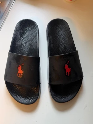 Svarta slides från Polo Ralph Lauren - Svarta sandaler/slides från Polo Ralph Lauren med röd logotyp framtill. Klassisk och enkel design med bred rem över foten och strukturerad sula för extra komfort. Perfekta för sommaren och chill dagar.