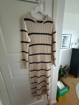 Randig långklänning från Pieces - Långärmad ribbad långklänning från Pieces i beige med svarta horisontella ränder. Klänningen har en rund halsringning och är tillverkad i ett mjukt stickat material som passar perfekt för kyligare dagar.