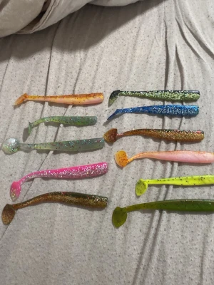 Färgglada mjuka fiskedrag helt oanvända - Säljer ett gäng helt oanvända mjuka fiskedrag i olika färger och former, perfekta för sportfiske. Drag i grönt, blått, rosa, gult, orange och glitter, tillverkade i mjukt plastmaterial. Passar till olika fiskesituationer och är lätta att använda.