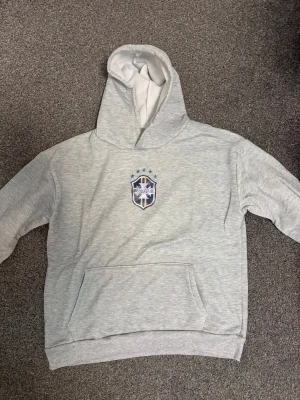 Grått mjukisset med tryck - Snyggt grått mjukisset med hoodie och byxor, båda med ett tryckt emblem framtill. Hoodien har magficka och byxorna har resår i midjan. Perfekt för en avslappnad stil. Materialet känns mjukt och bekvämt.