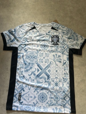 Mönstrad fotbollströja Louis Vuitton x Nike - Säljer en unik fotbollströja i samarbete mellan Louis Vuitton och Nike. Tröjan är vit med ljusblått mönster inspirerat av kakel, svarta sidopaneler och detaljer. Märkesloggor på bröstet och etikett nertill. Perfekt för dig som vill sticka ut på planen.