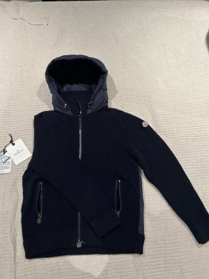 Moncler jacka  - Snygg mörkblå stickad jacka från Moncler med huva och dragkedja framtill. Jackan har två fickor med dragkedja och Moncler-logga på ärmen. Perfekt för kyliga dagar och har en ribbad struktur för extra stil.