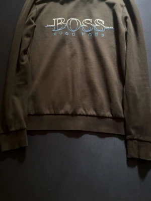 Svart sweatshirt från Hugo Boss - Snygg svart sweatshirt från Hugo Boss med stort BOSS-tryck och blå detaljer på bröstet. Tröjan har rund halsringning, långa ärmar och ribbade muddar. Perfekt för en avslappnad och trendig stil. Passar xs